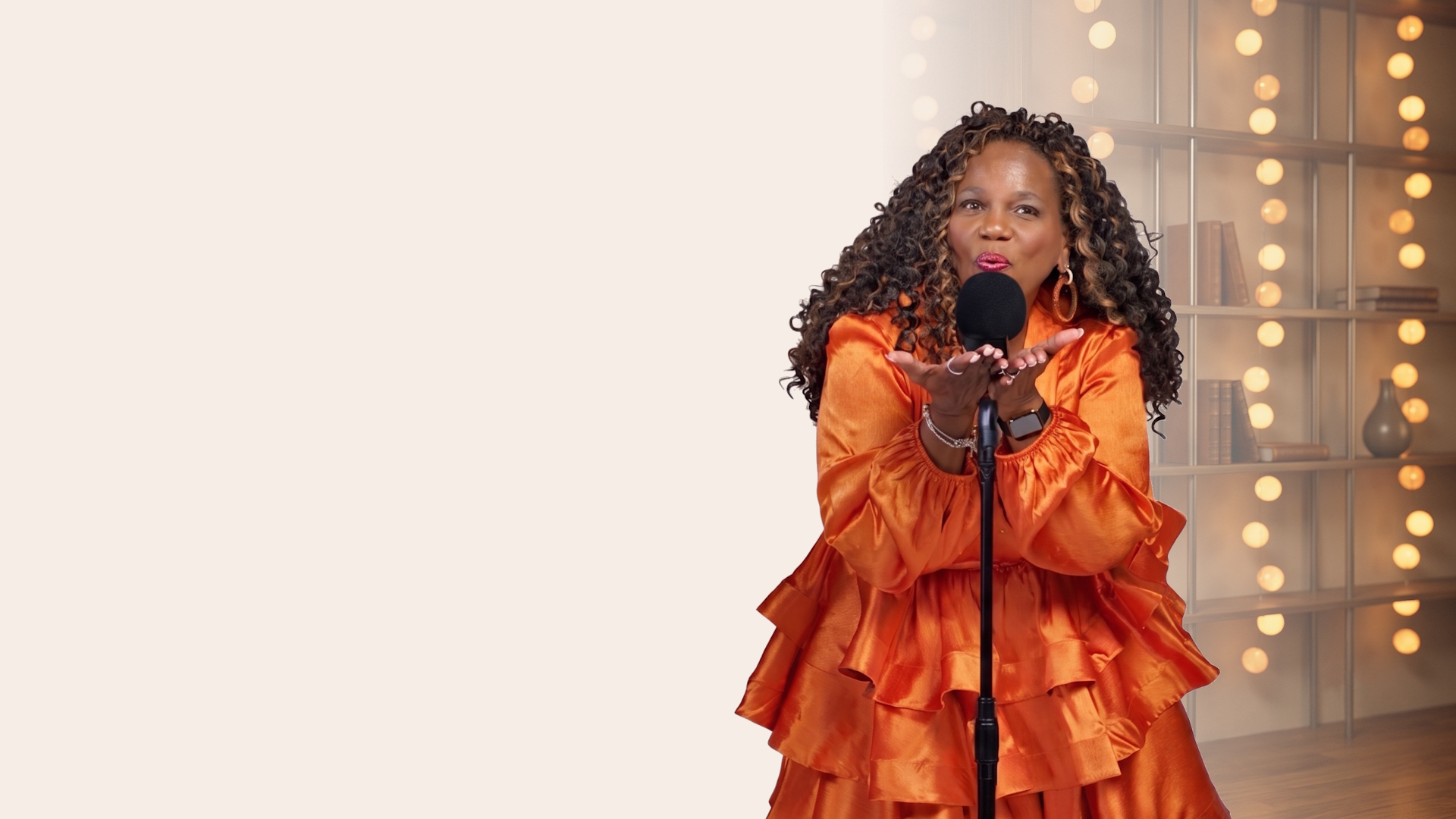 Lisha Lewis blowing a kiss toward a studio microphone, warm string lights glowing behind her